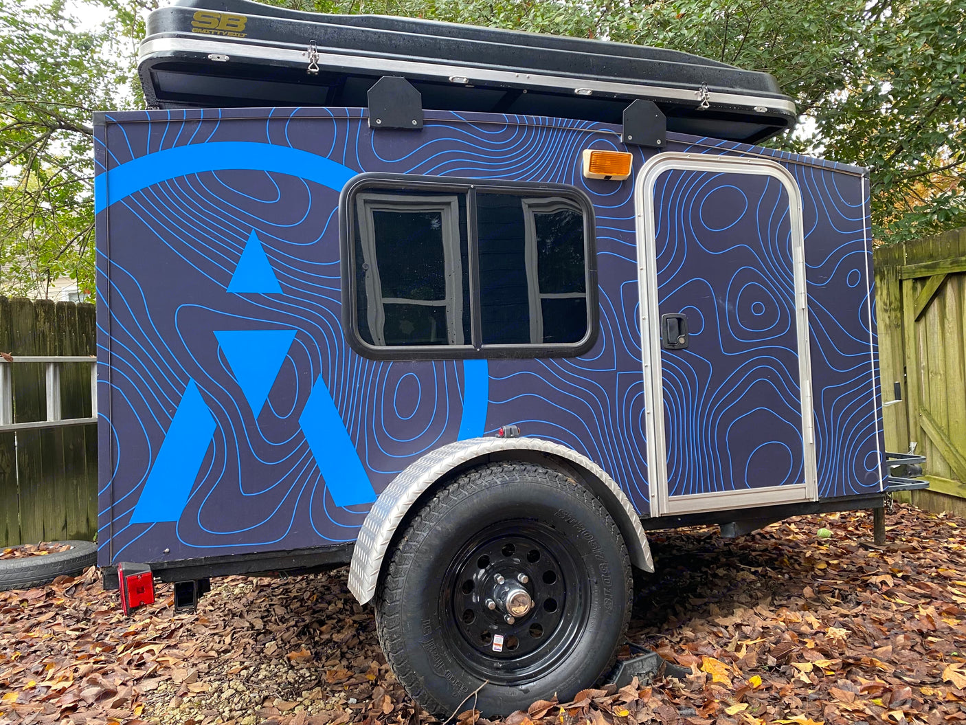Explore with this dark blue custom camper trailer, featuring a roof-top tent & unique topo-map graphics. Great for an RV rental!
