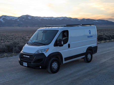 A white Vander RV rental van on a dirt road, backed by snow-capped mountains under a sunset sky.