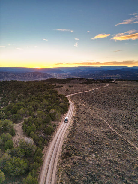 Experience an RV rental adventure! A white camper van travels a scenic dirt road through scrubland under a vibrant sunset.