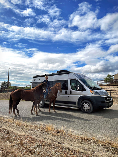 RV rental adventure: A person on horseback with a second horse pauses next to a camper van under a blue sky.