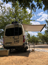 A tan Winnebago Revel RV rental parked at a dirt campsite, with an extended awning, table, and cooler. Perfect for glamping!