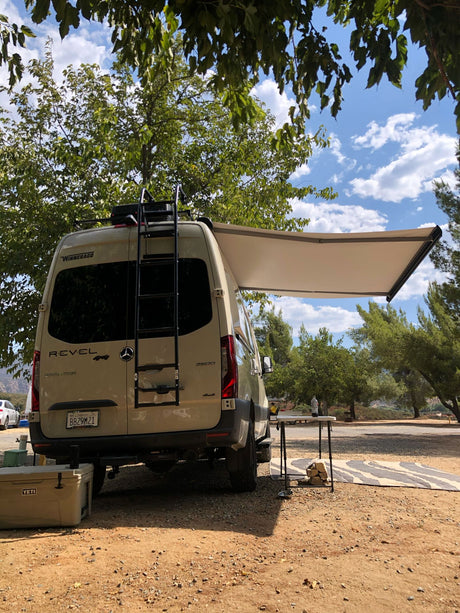 A tan Winnebago Revel RV rental parked at a dirt campsite, with an extended awning, table, and cooler. Perfect for glamping!