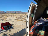 RV rental desert camping with outdoor cooking setup and van interior showing a bed, mountains in the background.