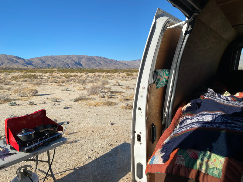 RV rental desert camping with outdoor cooking setup and van interior showing a bed, mountains in the background. — photo 7
