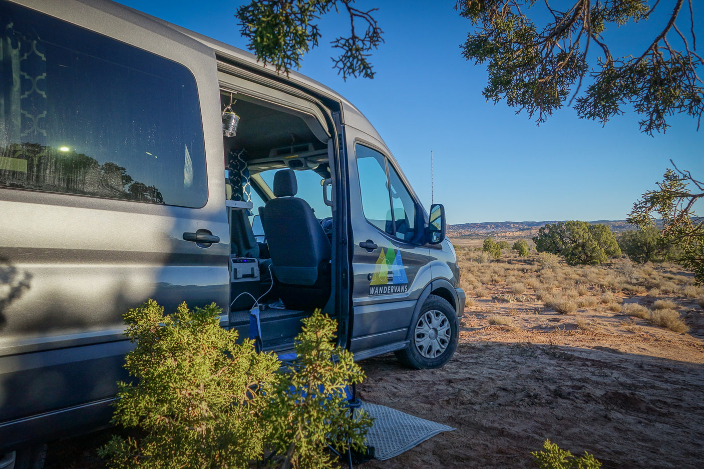 A grey Wandervans RV rental is parked in an arid desert landscape with its side door open, ready for adventure.