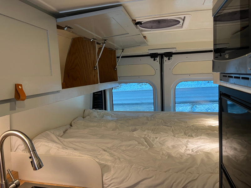 Inside a cozy rv rental: bed, overhead cabinets, kitchen faucet, and back windows with snowy views. Perfect compact living. — photo 11