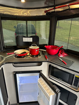 Cozy RV rental kitchen features stove, microwave, open fridge, and red cooking utensils, with a rainy outdoor view.