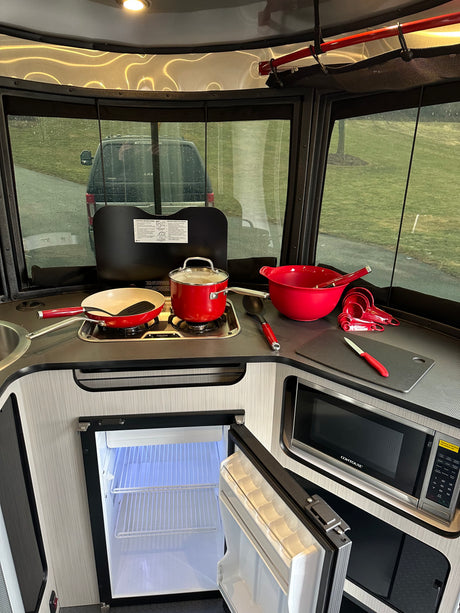 Cozy RV rental kitchen features stove, microwave, open fridge, and red cooking utensils, with a rainy outdoor view.