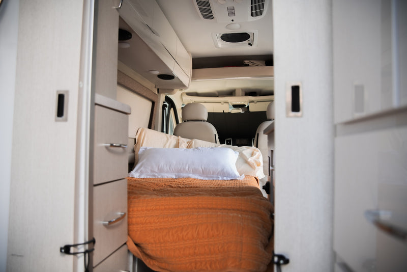 RV rental's inviting interior: a comfortable bed with an orange blanket & pillows, flanked by storage, leading to the front cabin. — photo 20