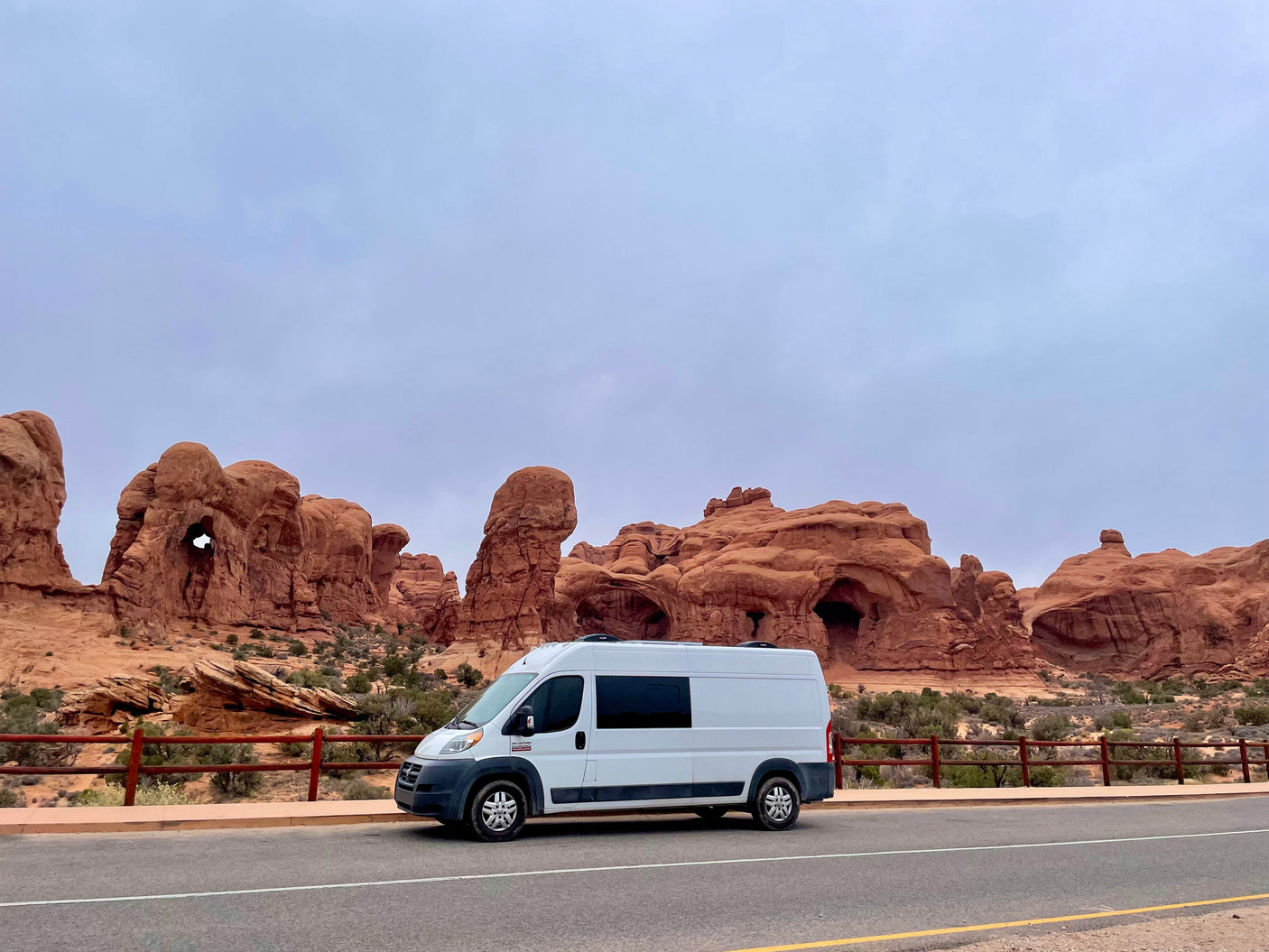 RV rental camper van parked amidst stunning red rock formations and natural arches in a desert landscape under an overcast sky.