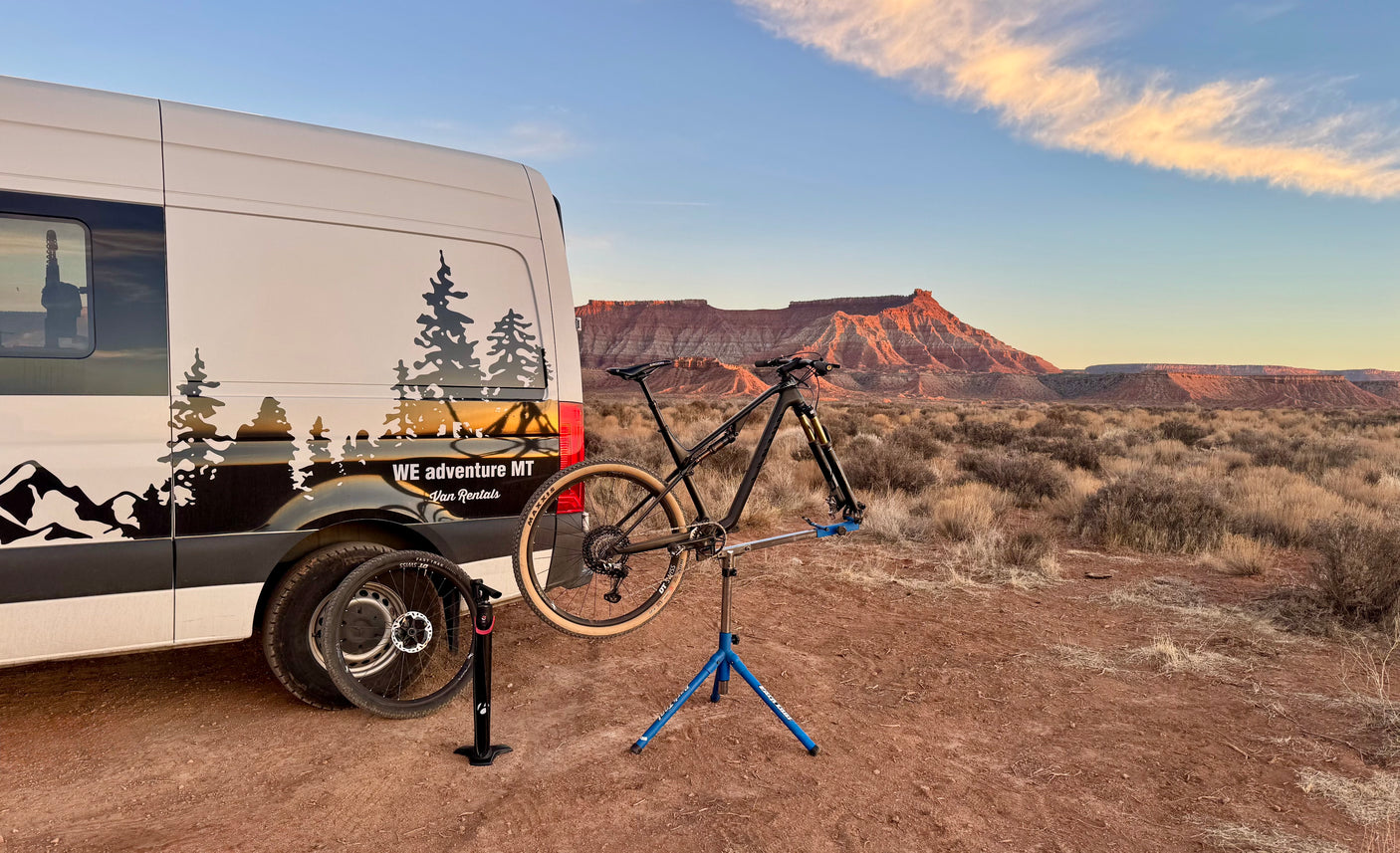 An RV rental van and mountain bike being serviced on a stand, set against a desert sunset.