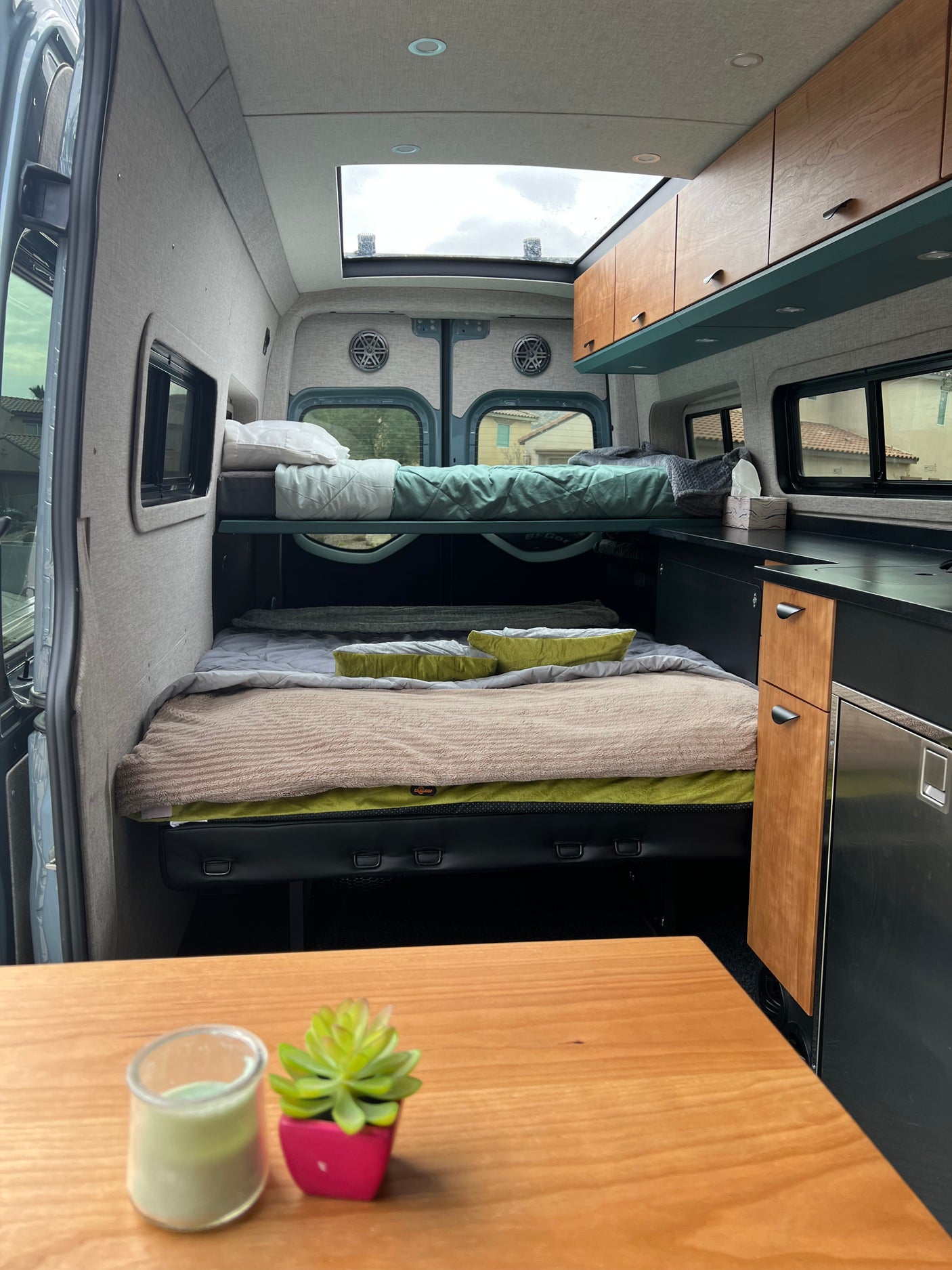 RV rental interior view: bunk beds, lower bed, compact kitchen with fridge/cabinets, and bright skylight above.