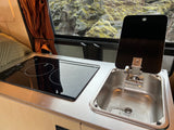 Compact RV rental kitchen features a running sink, induction cooktop, and a window looking out to mossy rocks.