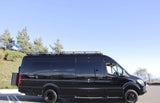 A sleek black Mercedes-Benz Sprinter camper van for rv rental, featuring off-road tires & a roof rack, parked outdoors.