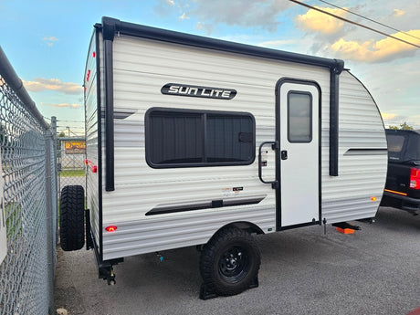White and gray Sun Lite travel trailer, a compact camper for RV rental, parked by a chain-link fence.