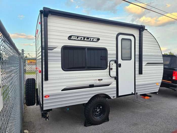White and gray Sun Lite travel trailer, a compact camper for RV rental, parked by a chain-link fence.