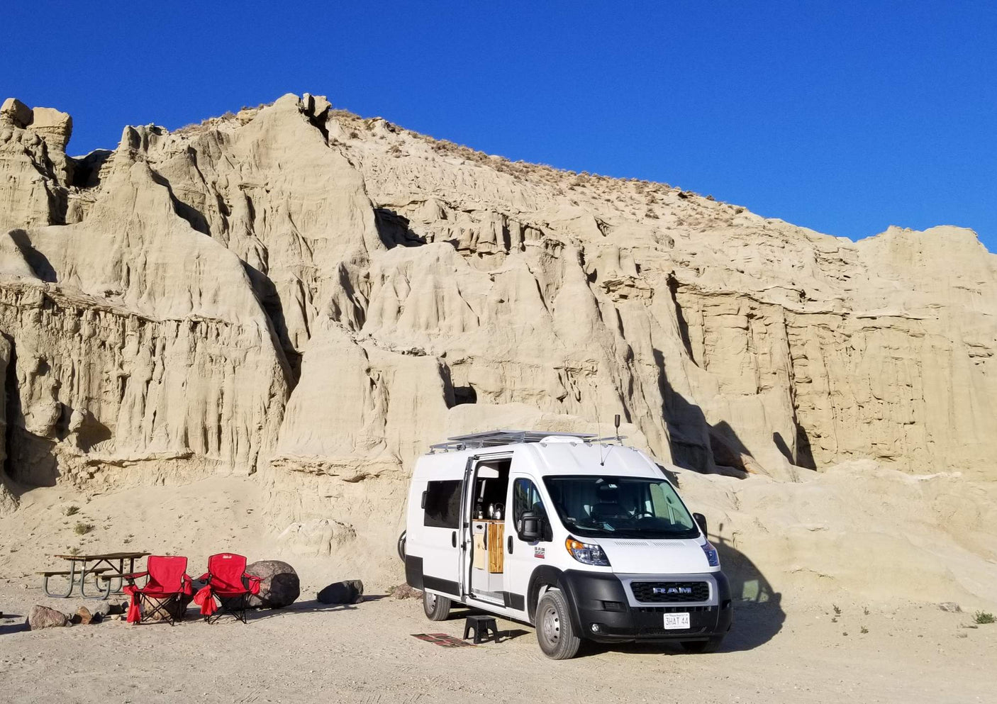 White RV rental van parked against dramatic, sculpted desert badlands. A picnic table and red chairs are set up for camping.