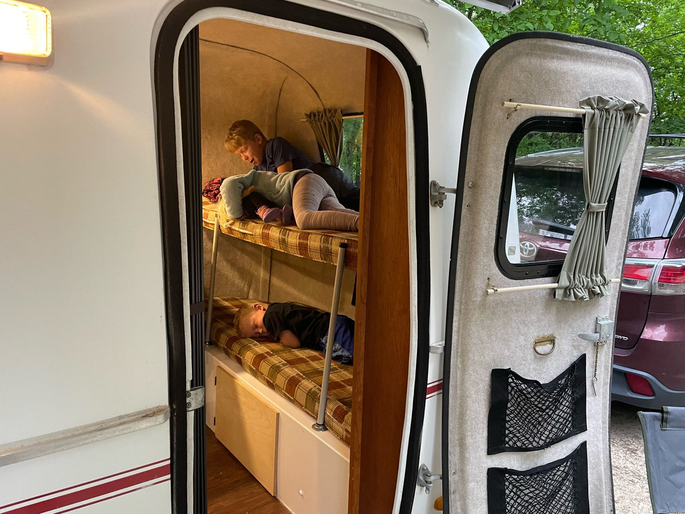 Inside an RV rental, two children are on bunk beds. The top bunk holds a child playing with a doll, while the bottom has a sleeping child. Compact and cozy camper.