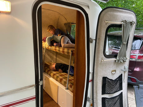 Inside an RV rental, two children are on bunk beds. The top bunk holds a child playing with a doll, while the bottom has a sleeping child. Compact and cozy camper.
