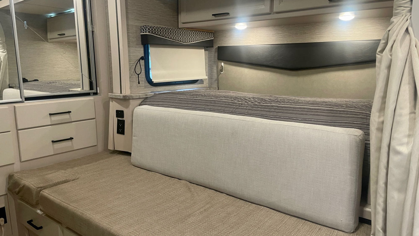 Inside a bright RV rental: view of a bedroom with bed, built-in mirrored storage, seating, and window with blind.