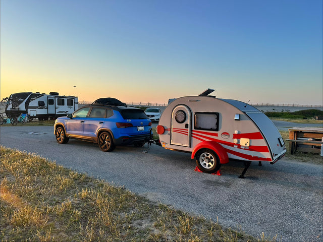 Blue SUV towing a T@G teardrop rv rental at a beachside campground during sunset. Another large RV is parked nearby.|A sleek white T@G teardrop trailer with red stripes