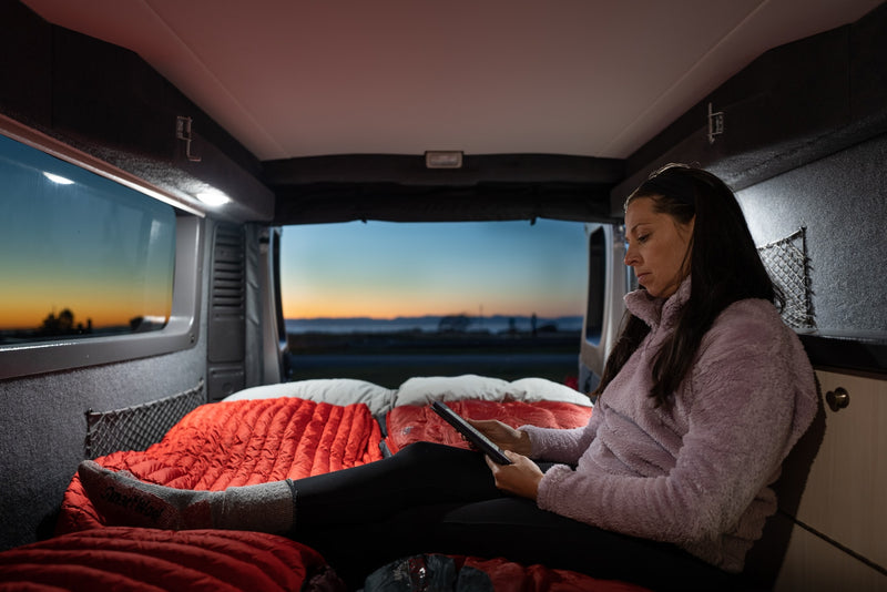 A woman relaxes with a tablet on a cozy bed inside an RV rental, enjoying comfort and a scenic sunset view. Perfect for travel. — photo 14