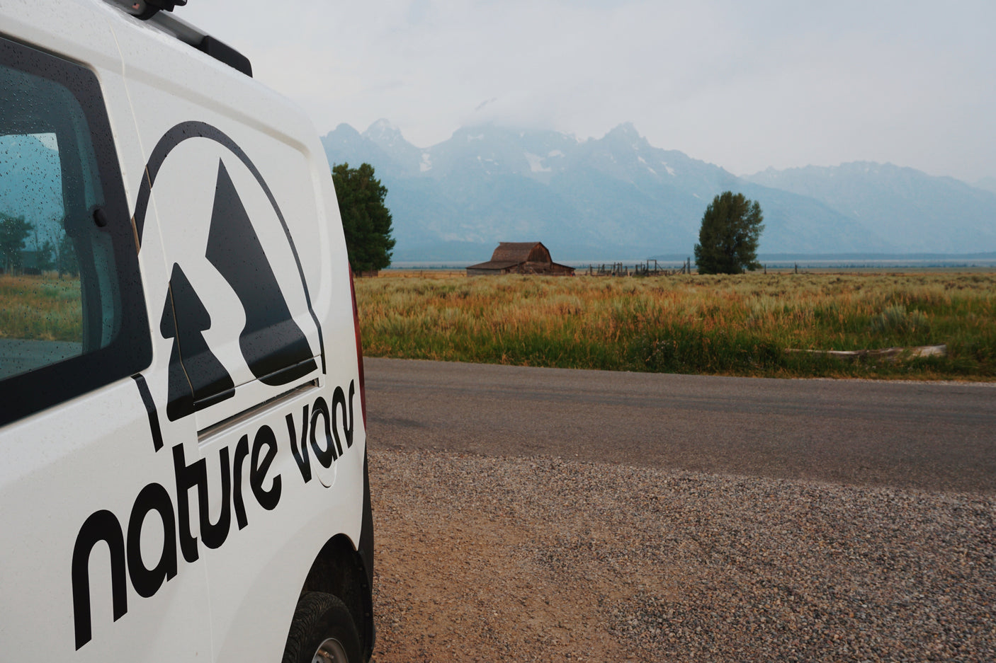 Explore with an RV rental! A nature van parked by a road, with scenic mountains, fields, and a historic barn in the background.
