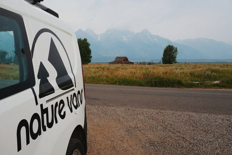 Explore with an RV rental! A nature van parked by a road, with scenic mountains, fields, and a historic barn in the background.