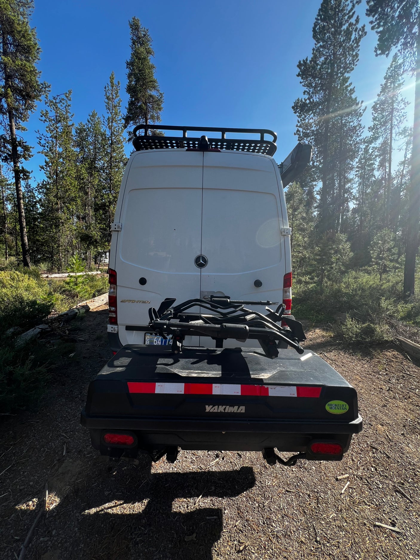 RV rental ready for adventure! Rear view of a white Sprinter van with a roof rack, bike rack, and Yakima cargo box in a forest.
