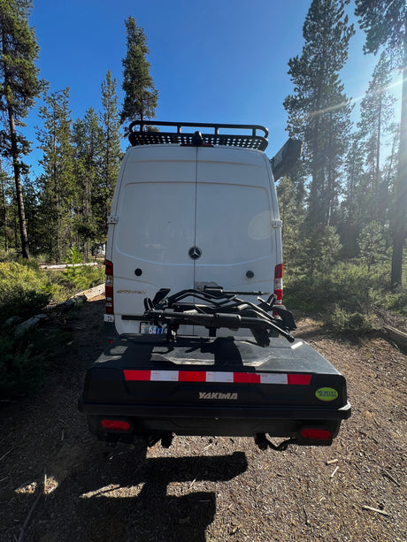 RV rental ready for adventure! Rear view of a white Sprinter van with a roof rack, bike rack, and Yakima cargo box in a forest.