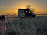 A serene sunset over a beige RV rental parked roadside. Two young people walk through golden tall grass with mountains in the distance.