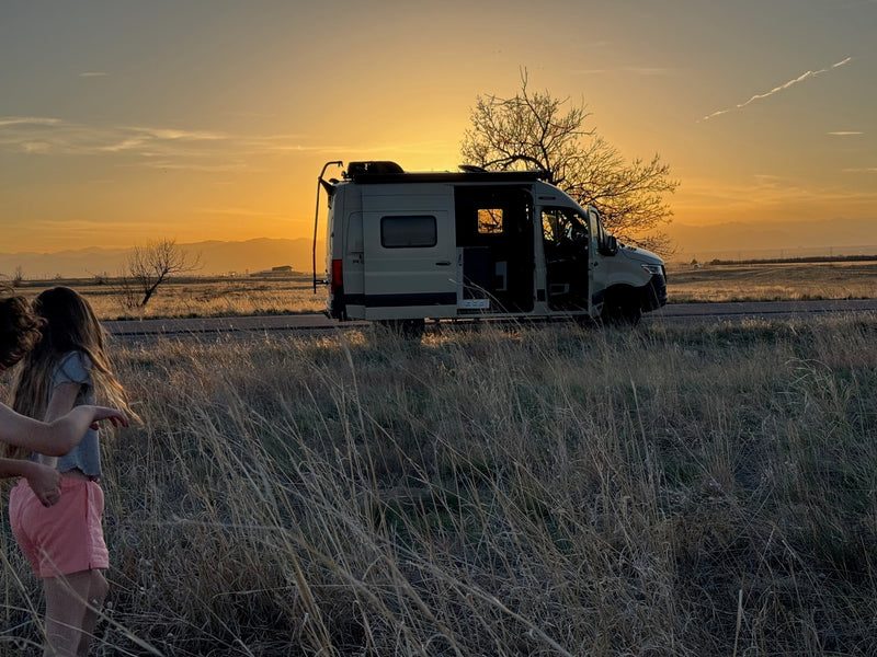 A serene sunset over a beige RV rental parked roadside. Two young people walk through golden tall grass with mountains in the distance. — photo 8