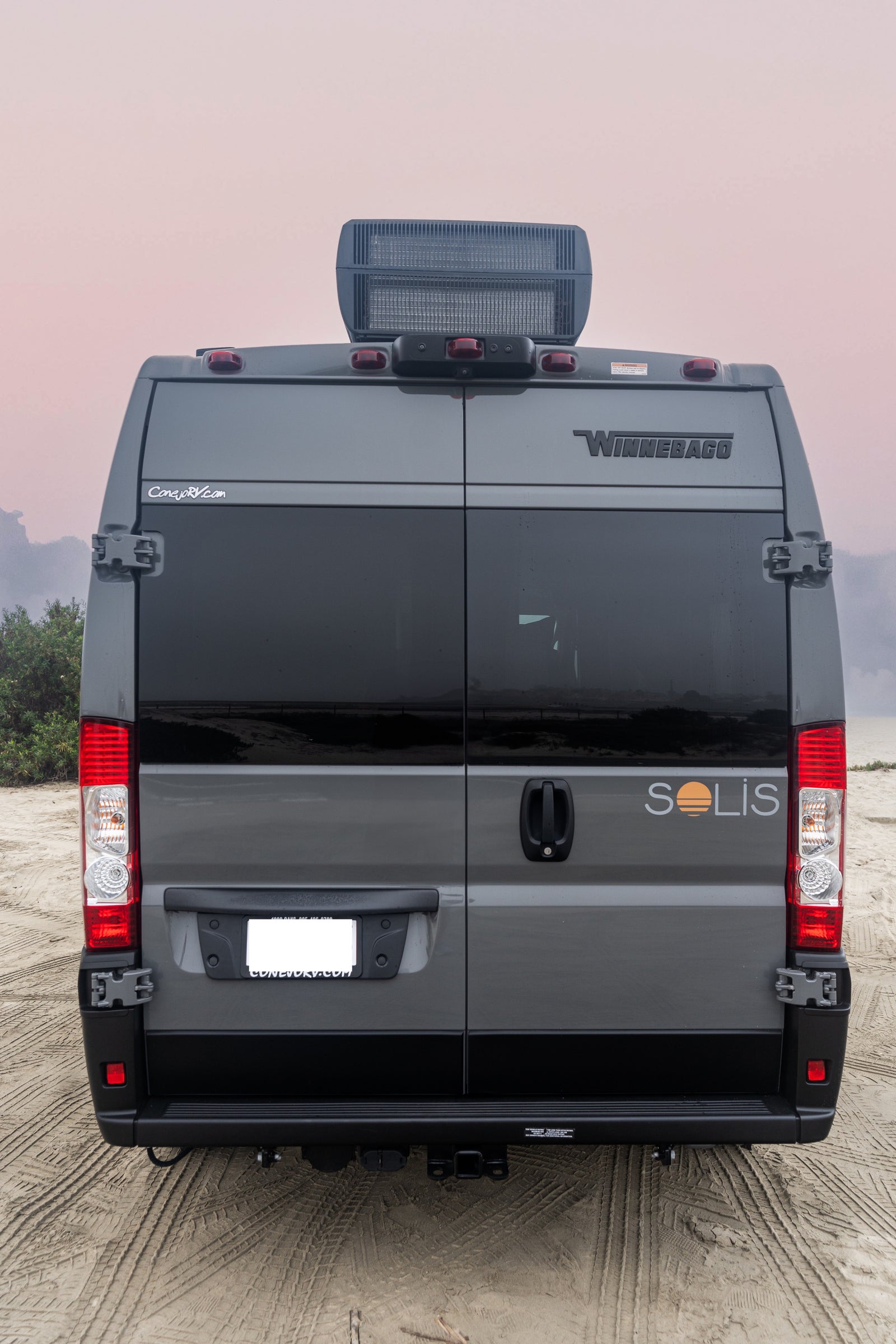 Rear view of a gray Winnebago Solis rv rental van on a sandy beach, with its distinct "Solis" sunset logo.