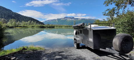 An rv rental, a compact travel trailer, perfectly situated by a calm mountain lake, reflecting snowy peaks & blue sky.