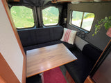 This RV rental interior features a functional dining area with black benches, a wooden table, and beautiful outdoor views.