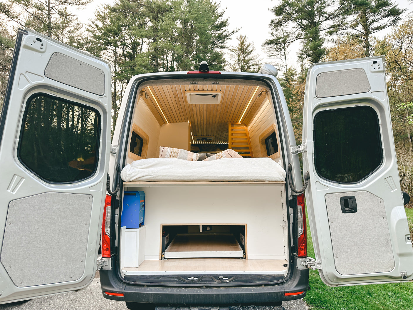 Rear view of a modern camper van conversion, ideal for your next RV rental trip. Features a bed, wood interior, & ample storage.