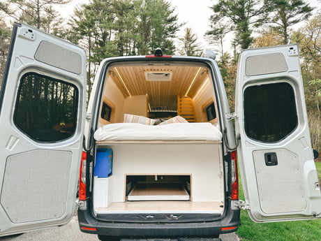 Rear view of a modern camper van conversion, ideal for your next RV rental trip. Features a bed, wood interior, & ample storage.