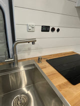 Modern RV rental kitchen: stainless steel sink, induction cooktop on wood counter, and wall controls on white paneling.