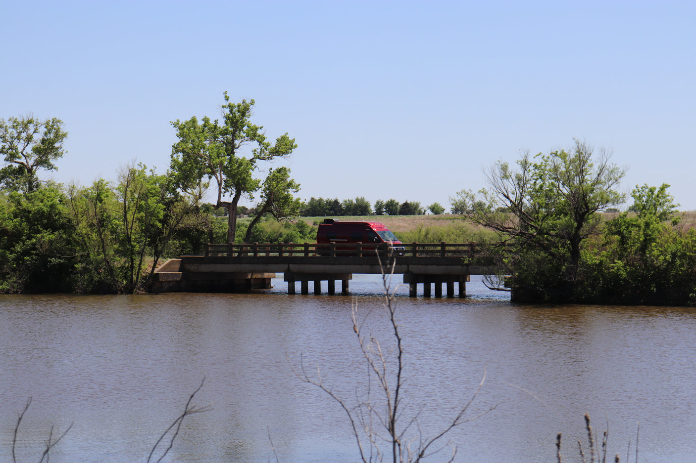 A red RV rental crosses a concrete bridge over a river, flanked by vibrant green trees under a clear sky.