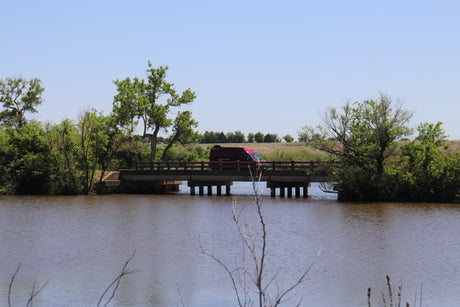 A red RV rental crosses a concrete bridge over a river, flanked by vibrant green trees under a clear sky.