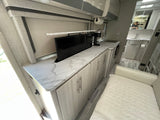 RV rental's modern kitchen: sleek marble-look counters, pop-up TV, light wood cabinets, and compact amenities.