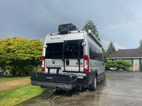 Rear view of a silver Thor Tellaro RV camper van on a wet driveway. A rainbow frames the sky. Looks like an RV rental ready to go!