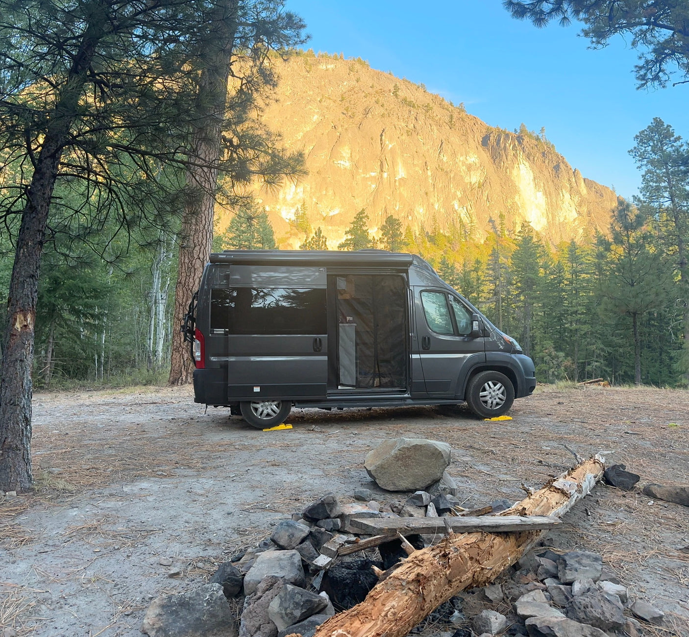 Rv rental camper van parked at a scenic campsite with a rocky mountain backdrop, towering pine trees, and a fire pit.