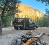 Rv rental camper van parked at a scenic campsite with a rocky mountain backdrop, towering pine trees, and a fire pit.