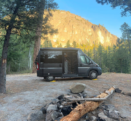 Rv rental camper van parked at a scenic campsite with a rocky mountain backdrop, towering pine trees, and a fire pit.