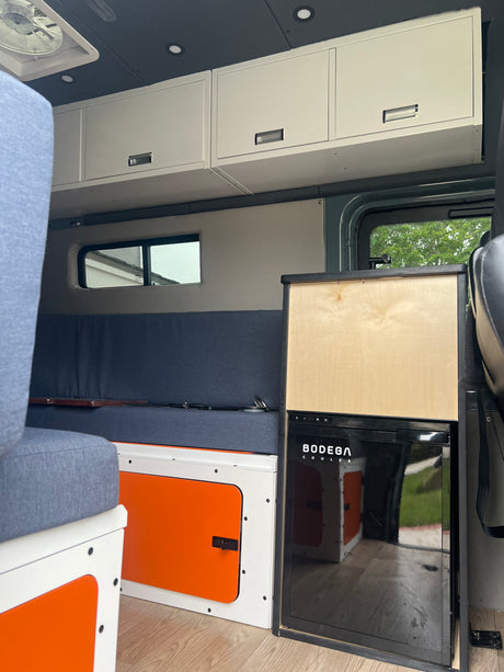 Inside this RV rental, find a compact interior with light wood flooring, white and orange cabinets, blue cushioned seating, and an integrated cooler.