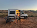 A man cooks dinner next to a white RV rental camper van at sunset in a desert landscape.