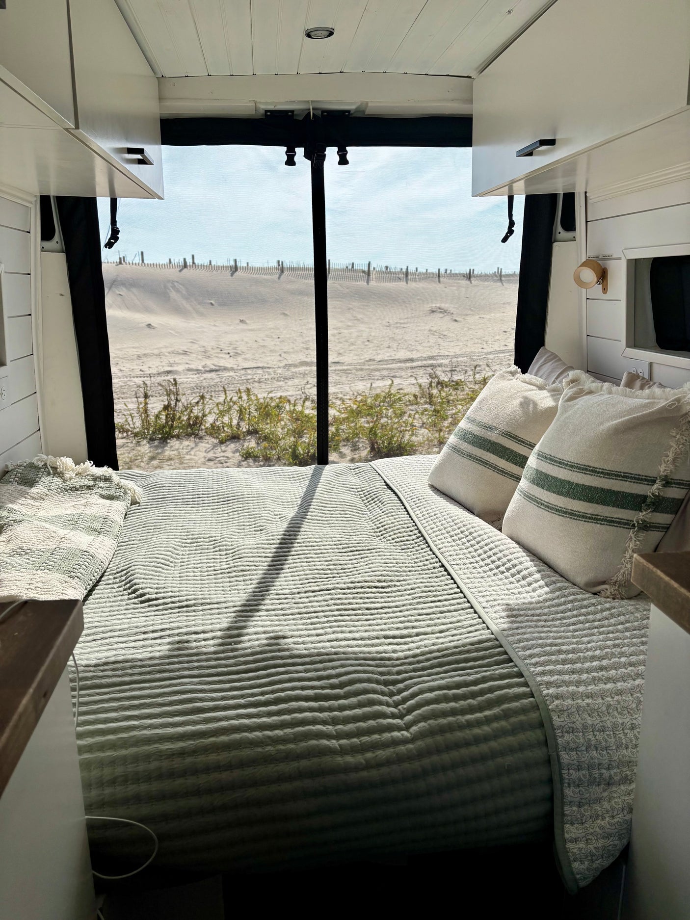 Inside a cozy RV rental: a bed facing a sandy beach view, perfect for van life adventures by the sea.