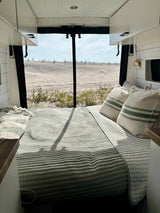 Inside a cozy RV rental: a bed facing a sandy beach view, perfect for van life adventures by the sea.