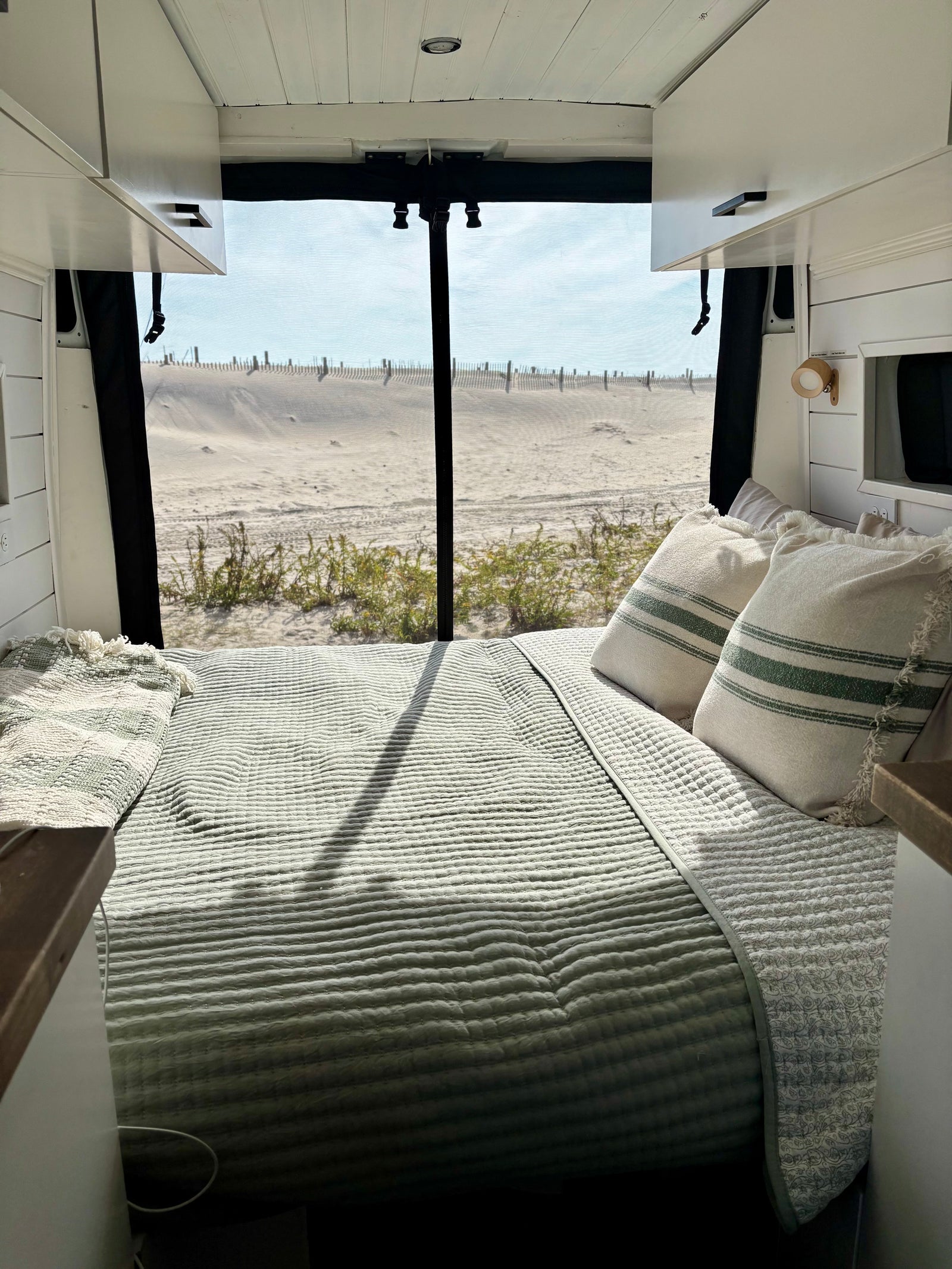 Inside a cozy RV rental: a bed facing a sandy beach view, perfect for van life adventures by the sea.